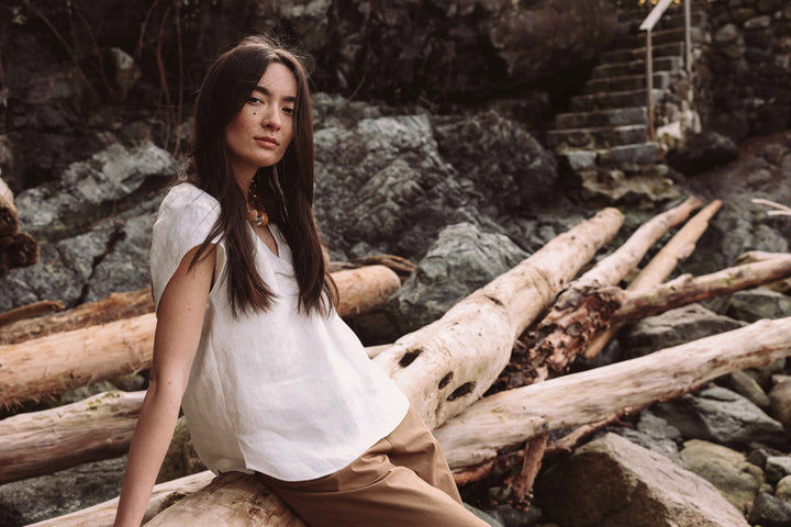 Woman wearing Georgia Blouse (white flowy blouse with a cap sleeve) sitting on driftwood